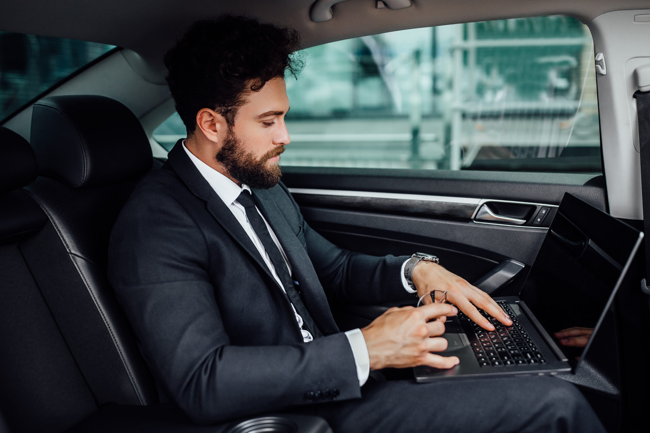 Professional in suit using laptop in car, showcasing car rentals in Delhi for corporate meetings.